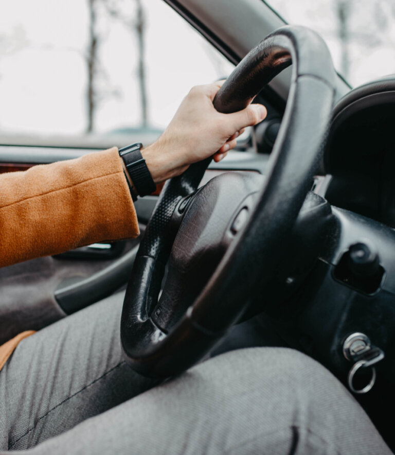 Close-up of a person’s hand on a steering wheel, wearing a brown coat and smartwatch